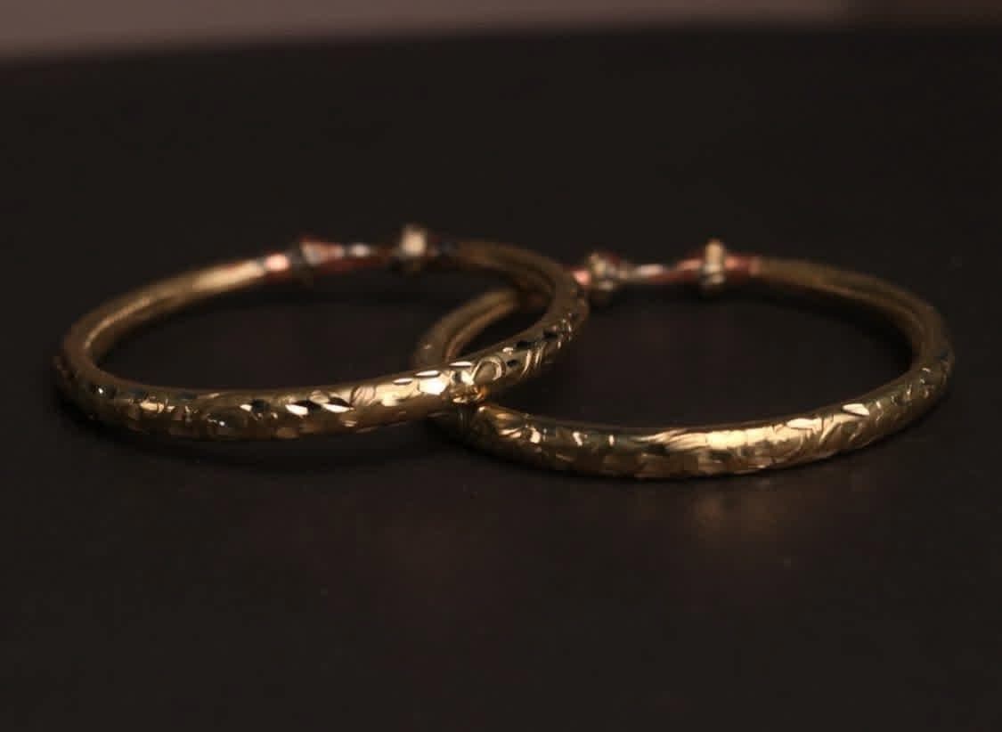 A pair of golden Pipe Bala bangles with an etched pattern, on a dark background.